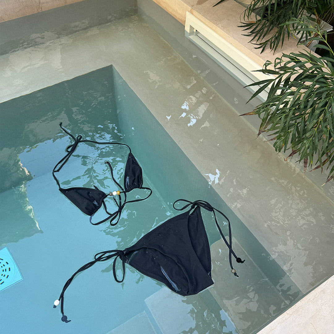 Black bikini XS/S with triangle top, chunky beads, and high-cut bottoms featuring side ties. Comes in a sleek black pouch, ideal for beach days.