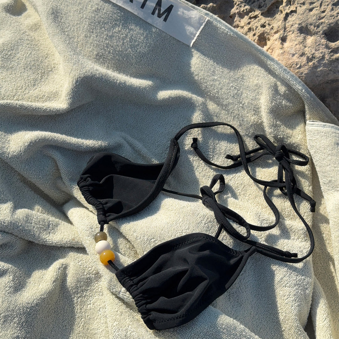 Black bikini with triangle top, earthy-tone beads, high-cut bottoms, side ties, and decorative straps, packed in a black drawstring pouch labeled 'Pilgrim', size M/L.