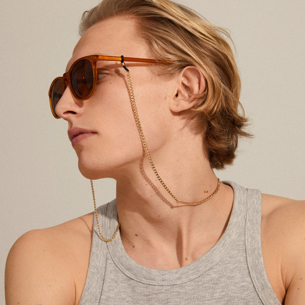 A man wearing sunglasses with the AJA curb chain for sunglasses gold-plated, showcasing its stylish, versatile design with black adjustable loops.