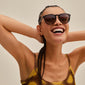 Woman smiling, wearing Pilgrim's SAHARA sunglasses in tortoise brown, showcasing trendy eyewear with UVA/UVB protection. Her hands are behind her head, radiating confidence.