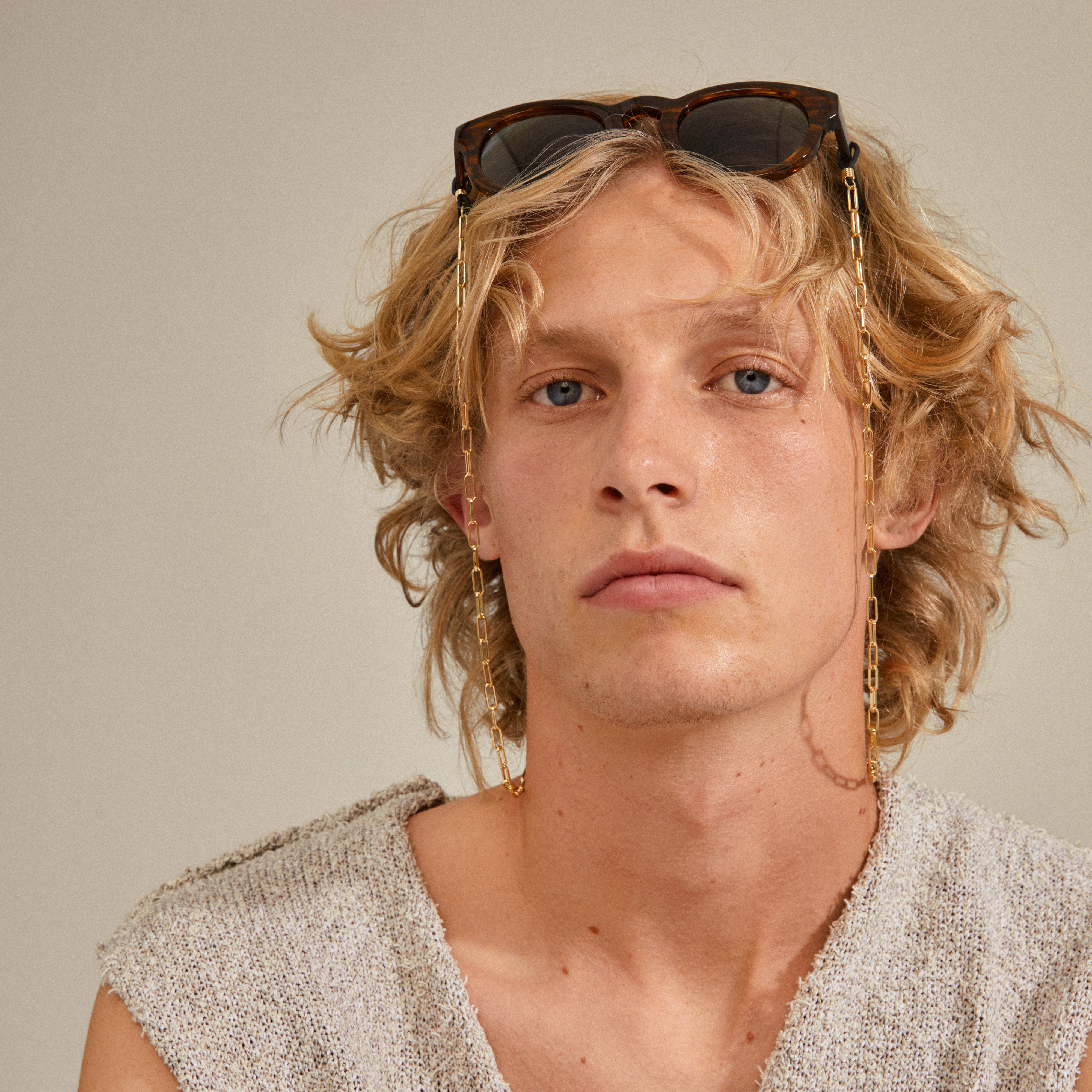 Close-up of a man with long blonde hair wearing sunglasses with the PAOLA gold-plated chain for sunglasses, showcasing its stylish cable-chain design.