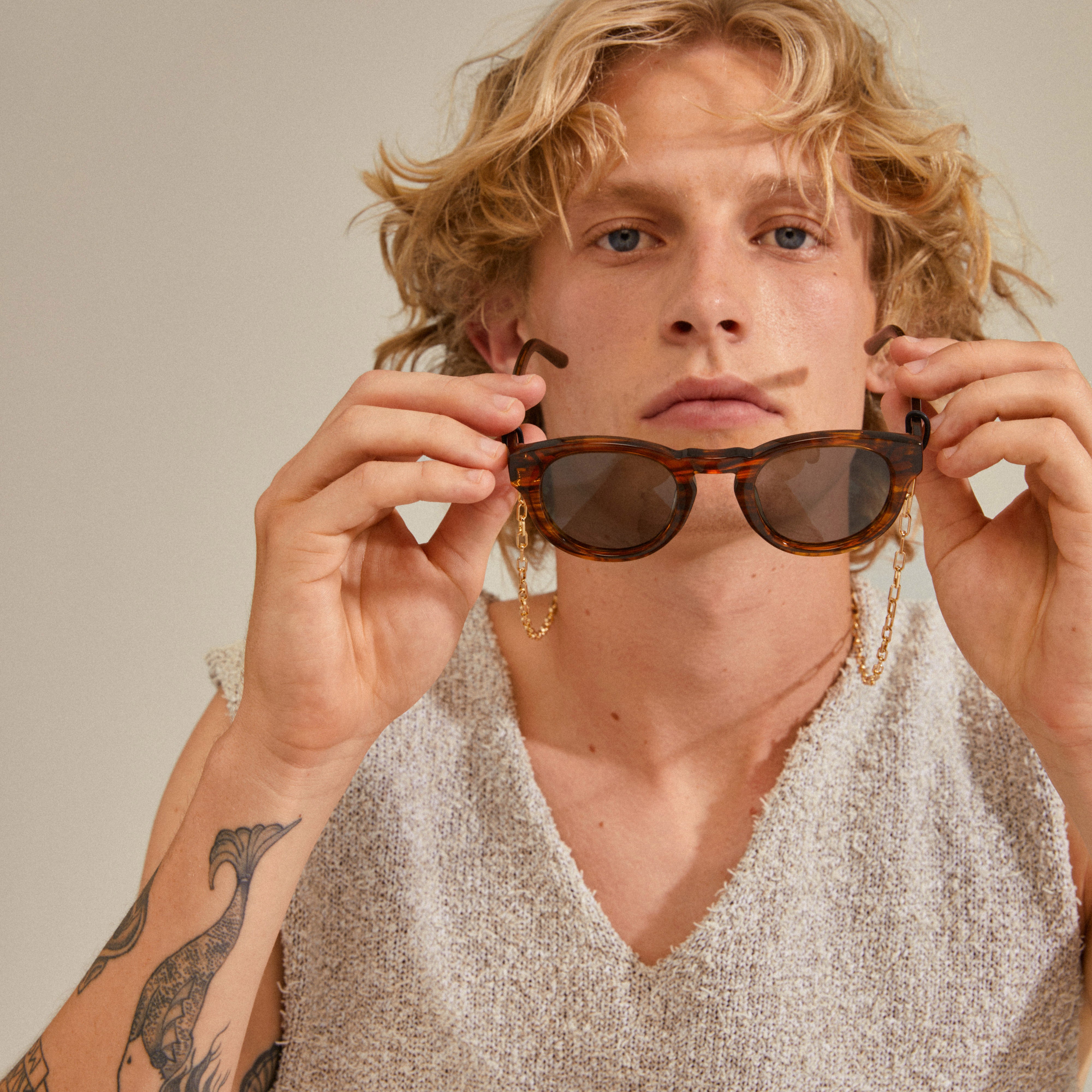 Man holding sunglasses with a gold-plated PAOLA chain from Pilgrim, showcasing its delicate cable-chain design attached with black rubber loops.