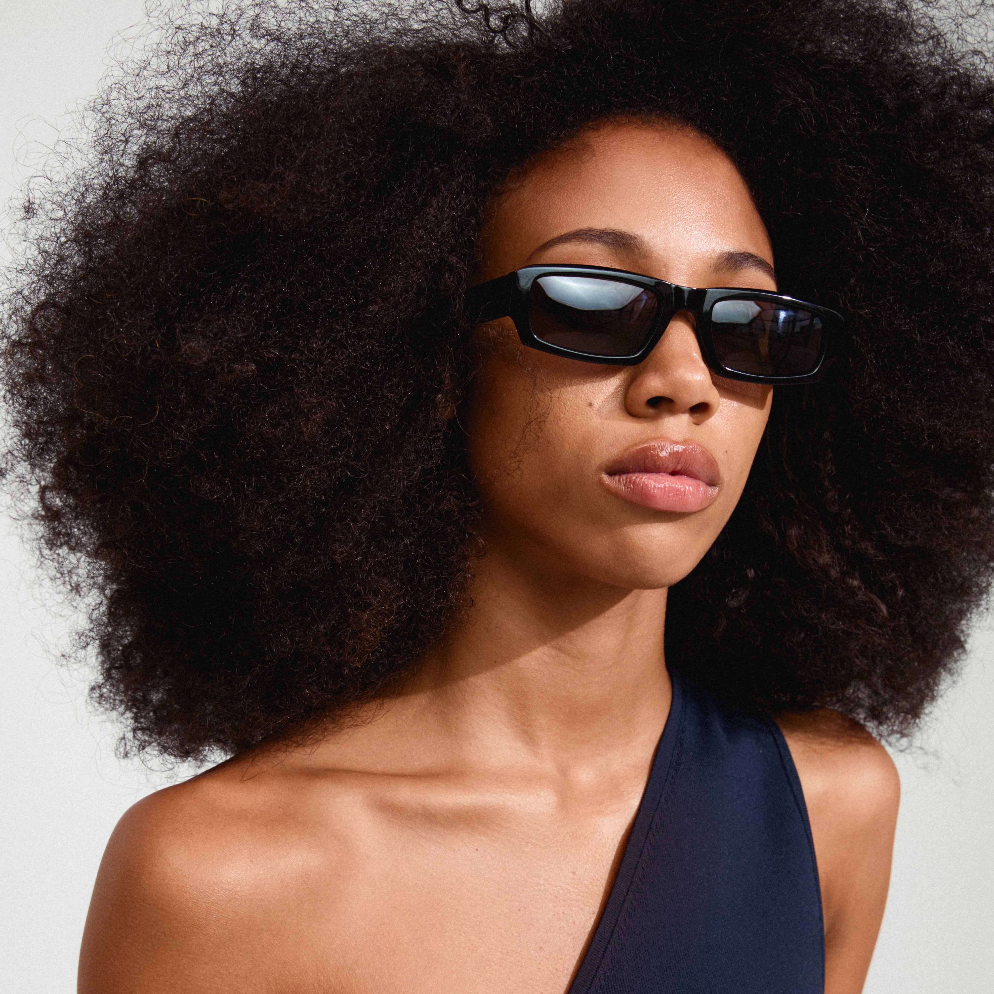 Woman with an afro wearing ALAYNA sunglasses featuring wide chunky temples, slim angular frames, embodying urban retro style.