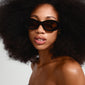 ALEXANDRIA sunglasses black feature a chunky eclipse-shaped frame and are worn by a woman with an afro, showcasing retro-inspired fashion.