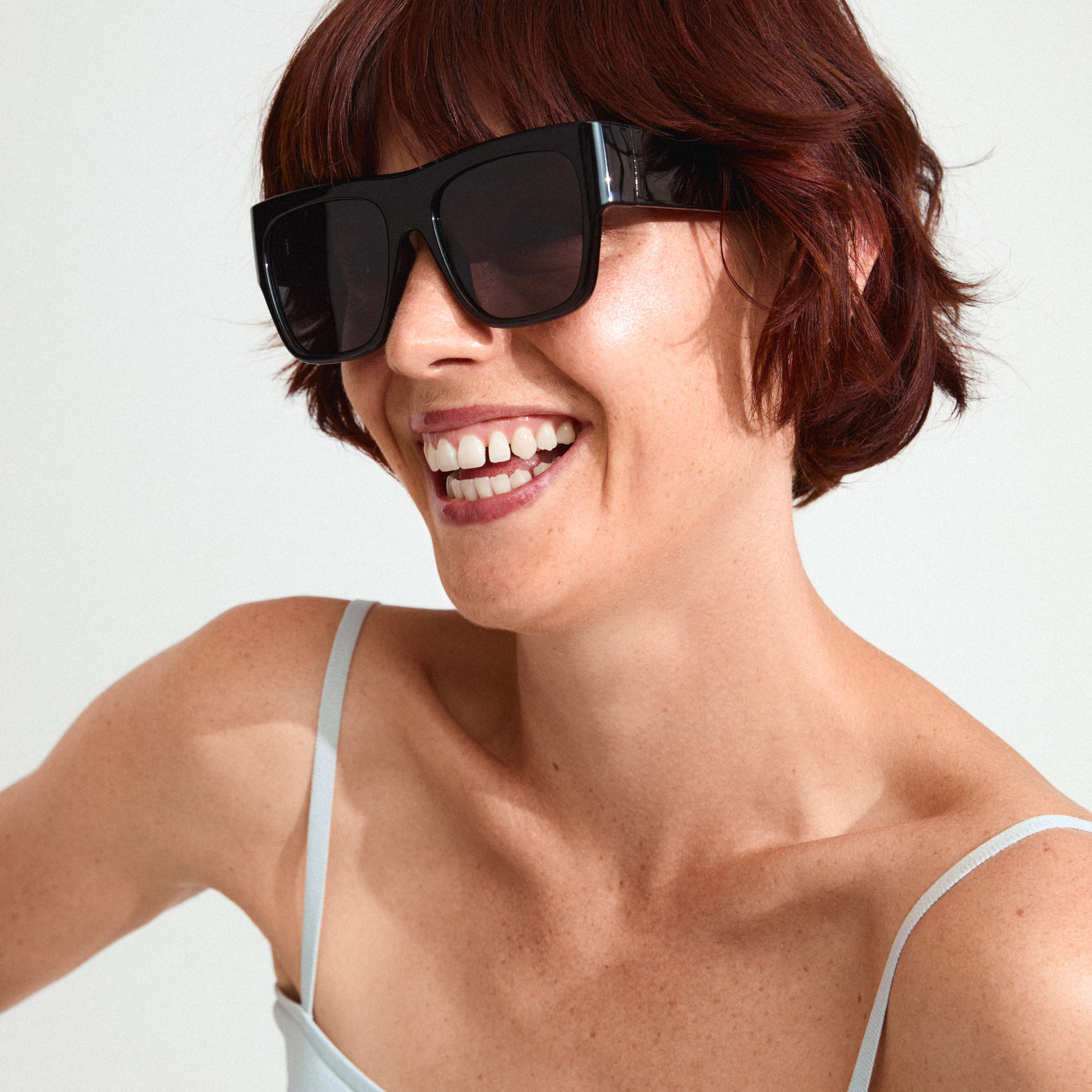 Smiling woman wearing ANIYAH sunglasses from Pilgrim, featuring oversized black frames with dark lenses, embodying chic runway fashion and movie-star allure.