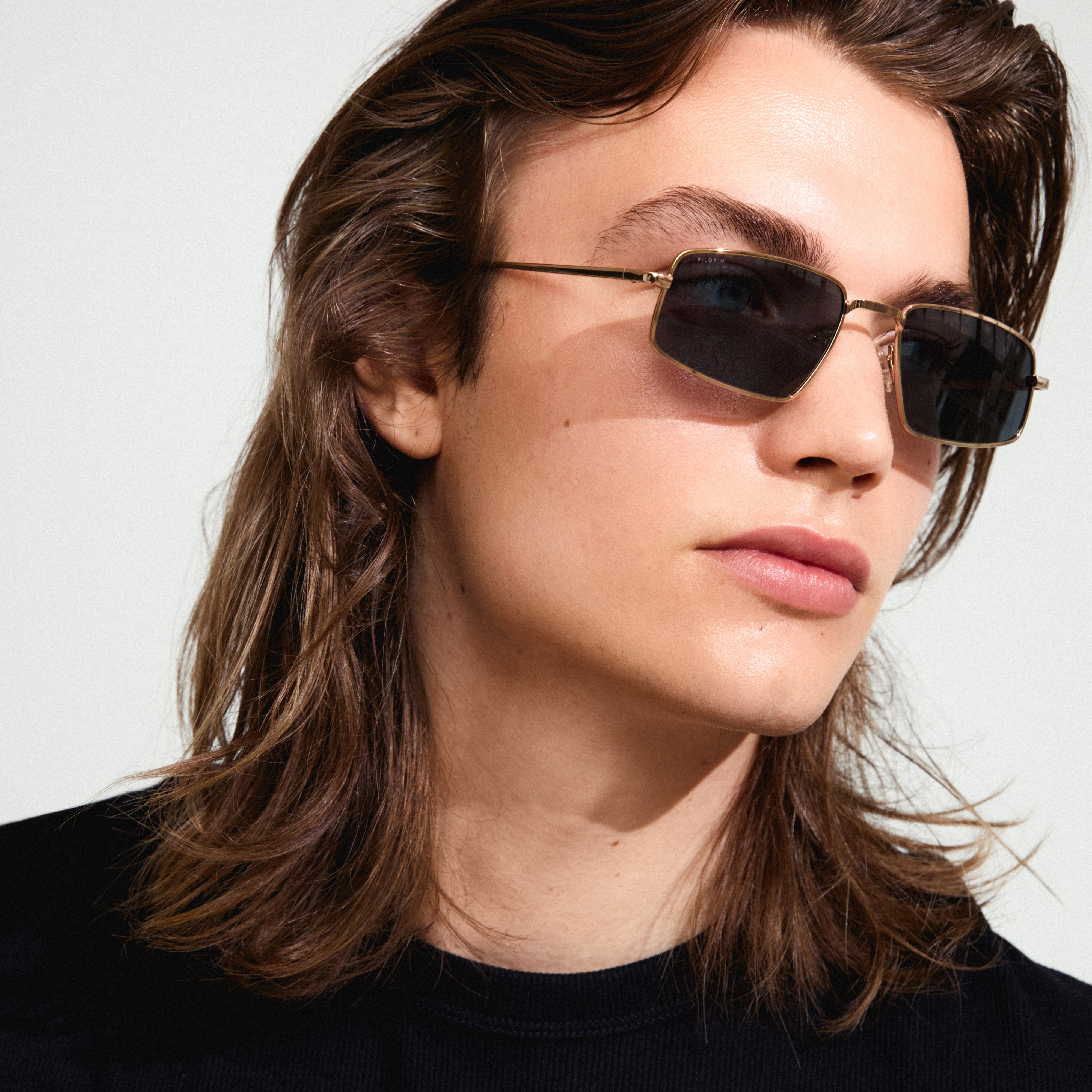 Man with long hair wearing AMIYAH sunglasses by Pilgrim, featuring edgy gold frames and smoke-colored lenses, perfect for adding attitude to any outfit.