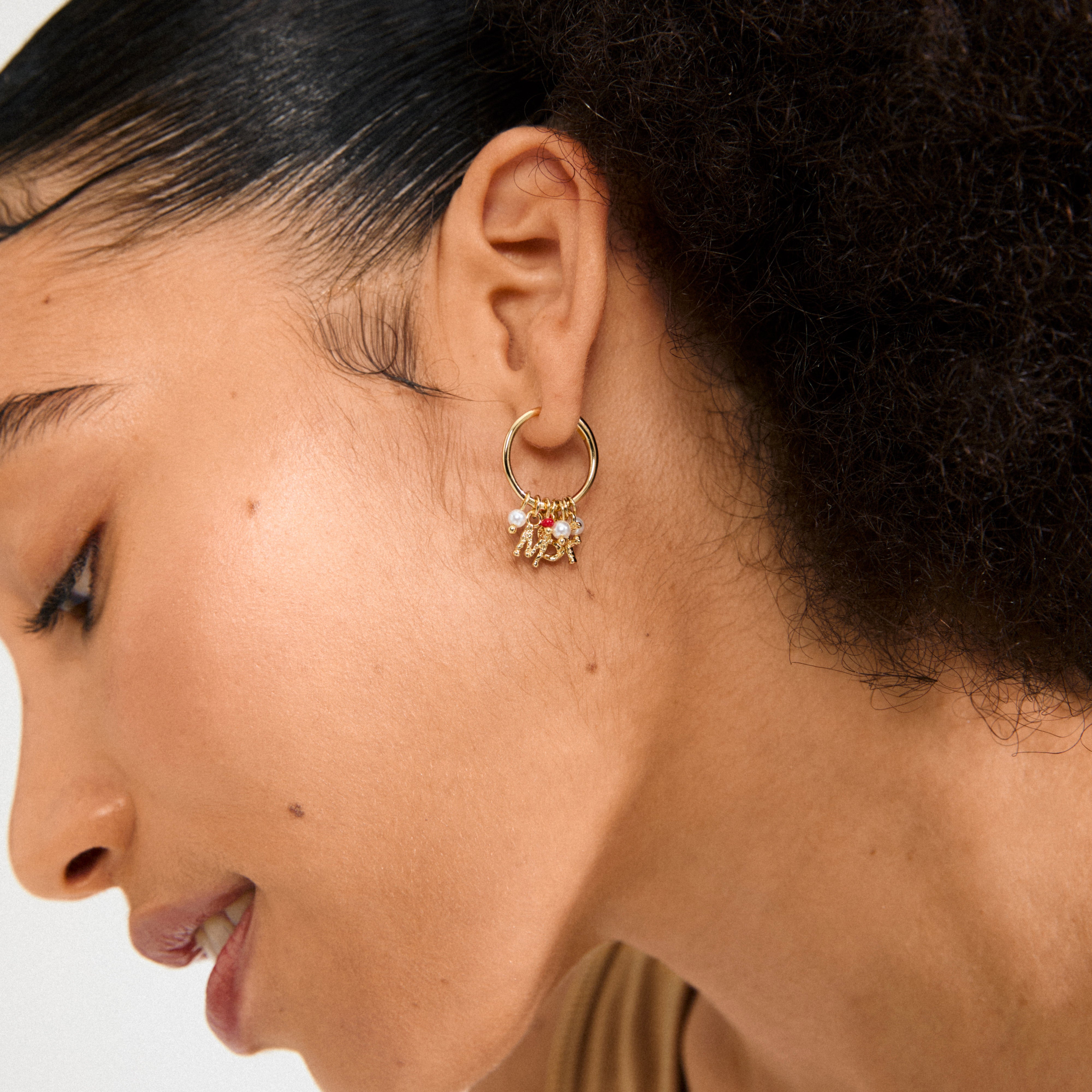 Close-up of MSF earrings gold-plated, featuring dangling charms with pearls, red bead, and gold trinkets on a person’s ear, supporting Doctors Without Borders.