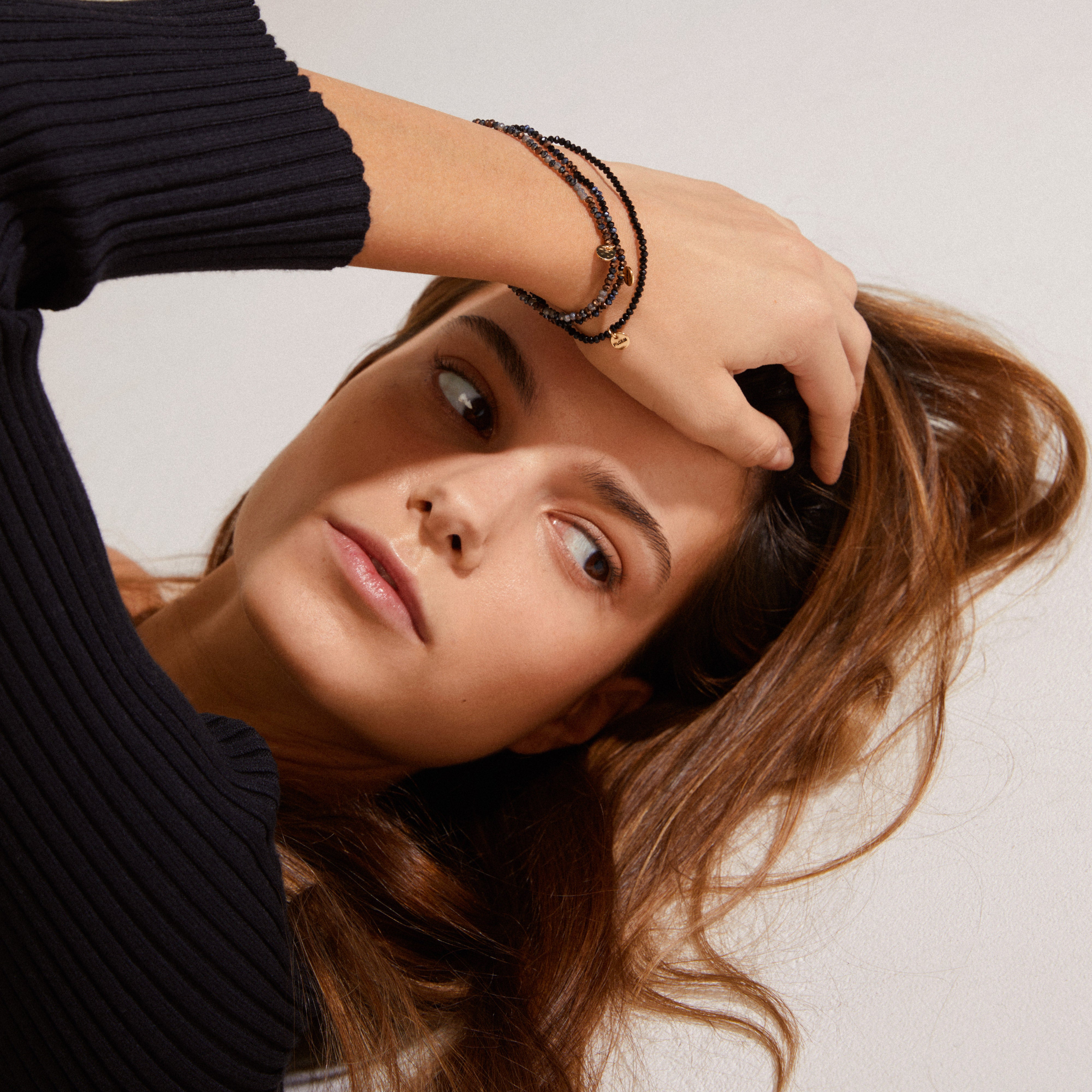 Young woman wearing the INDIGO 3-in-1 bracelet set, grey-black/gold-plated, with delicate beaded bracelets and small charms on her wrist, styled elegantly.
